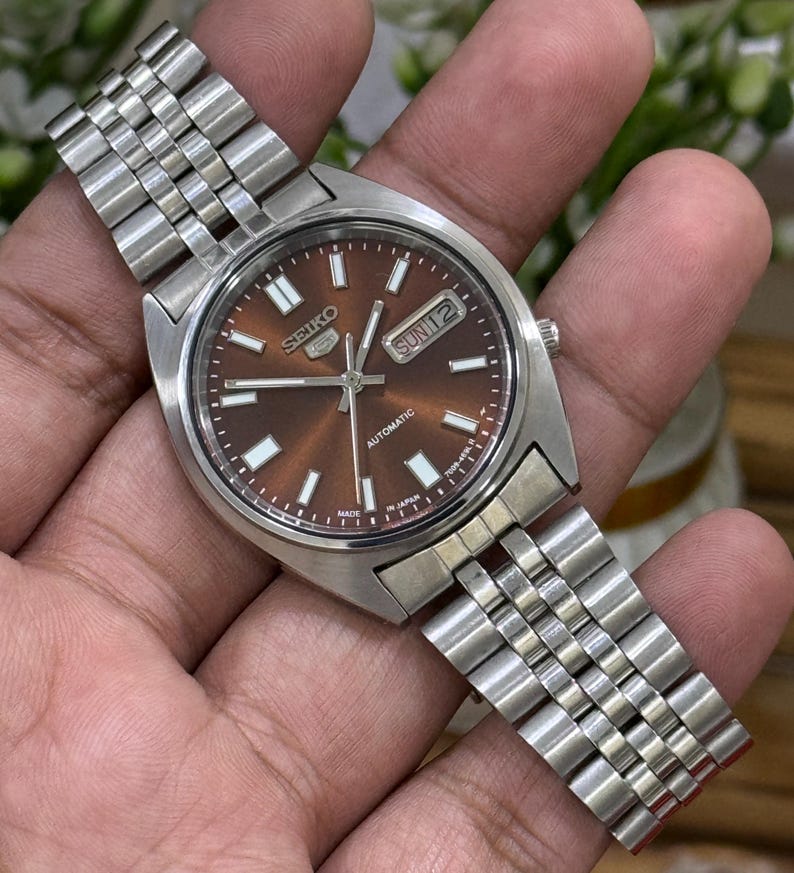 Może przedstawiać: Automatyczny zegarek Seiko 5 ze stali nierdzewnej z brązową tarczą. Zegarek posiada wyświetlacz dnia i daty oraz srebrną metalową bransoletę. Zegarek jest trzymany w dłoni.
