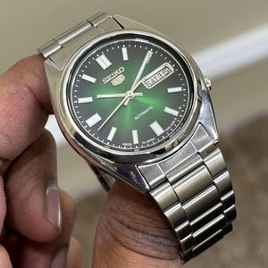 May include: A stainless steel Seiko 5 automatic watch with a green dial. The watch features a silver band, white hour markers, and a day-date display. The watch is held in a hand.