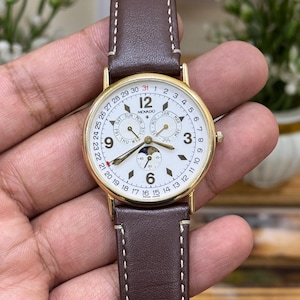May include: A gold Movado watch with a white face, displaying the date and moon phases. It has a brown leather strap with white stitching. The watch face shows the days of the week and months. The watch's diameter is approximately 3.5 cm.