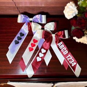 Può includere: Tre fiocchi decorativi in nastro viola, bianco e bordeaux, ciascuno con un diverso design a cuore. Un nastro ha il testo "ABCD 1234". I fiocchi sono disposti su una superficie di legno scuro.