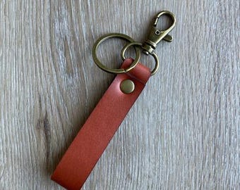May include: A leather keychain with a rust-colored strap and a brass-colored clasp and key ring. The strap is attached to the ring with a brass-colored rivet. The keychain is laying on a light brown wooden surface.