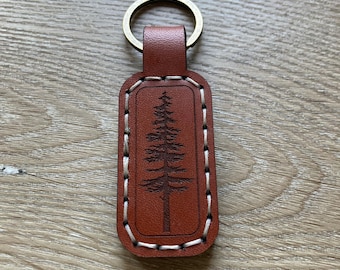 May include: A brown leather keychain with a gold-colored ring. The rectangular tag features a stitched border and a detailed tree design. The keychain is a practical accessory.