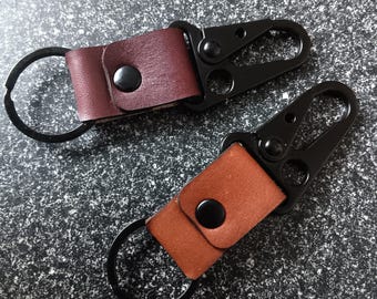 May include: Two leather keychains with black metal carabiner clips. One keychain has a burgundy leather strap, and the other has a tan leather strap. Each has a black metal ring and a black snap closure.