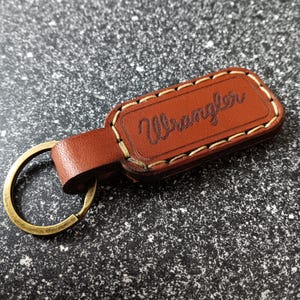 May include: Brown leather keychain with a stitched border and the word "Wrangler" embossed in white. The keychain has a brass key ring.