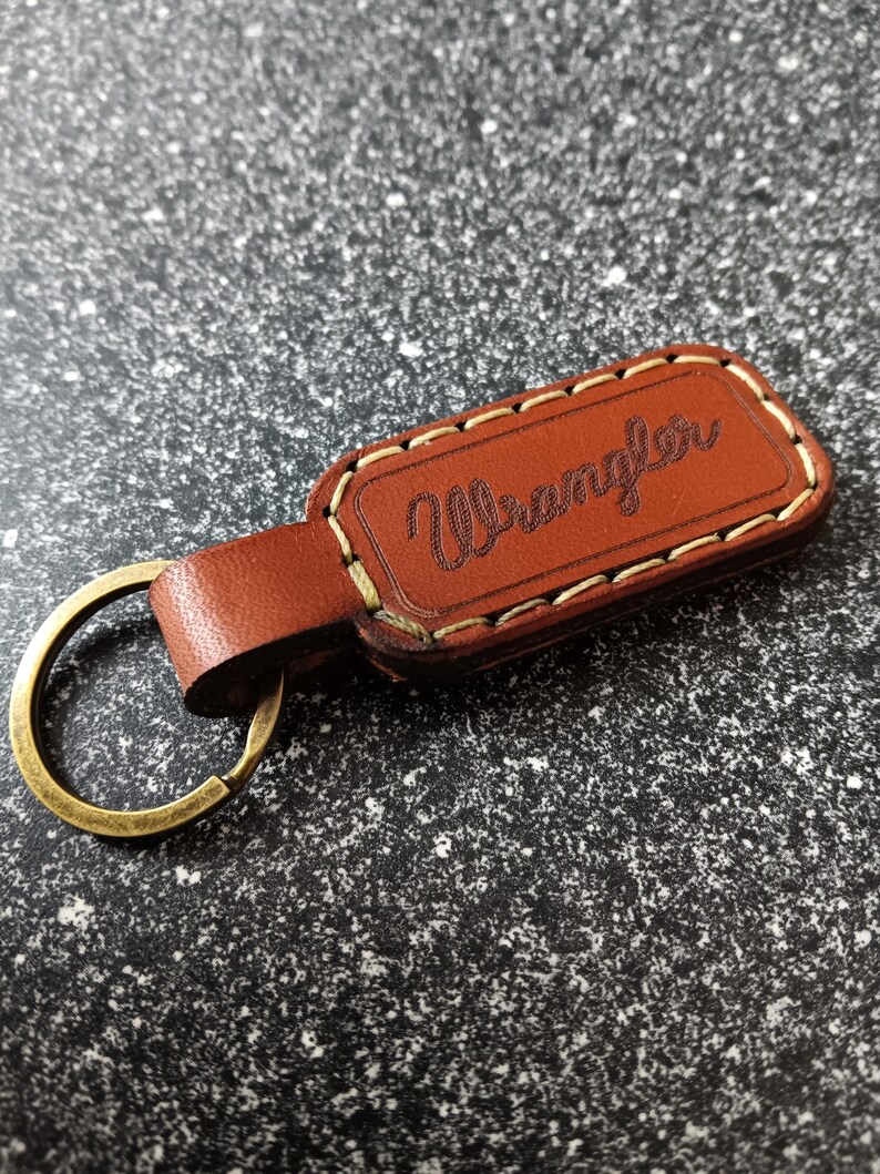 May include: Brown leather keychain with a stitched border and the word "Wrangler" embossed in white. The keychain has a brass key ring.