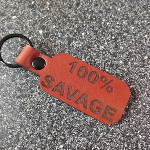 May include: A red leather keychain with the words "100% SAVAGE" engraved in the center. The keychain has a black metal ring and a black button. The leather is a reddish-brown color.
