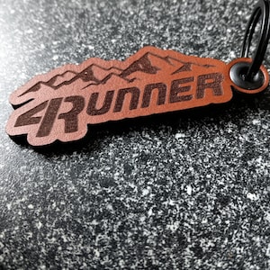 May include: Brown leather keychain with a mountain range design and the text "4Runner" engraved on it. The keychain has a black metal ring.