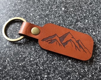 May include: A brown leather keychain with a mountain range design. The rectangular tag has a brass rivet and a gold-colored ring. The mountain graphic is etched into the leather.