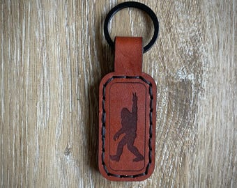 May include: A brown leather keychain featuring a silhouette of Bigfoot giving a peace sign. The keychain has a black key ring and black stitching around the edges. The background is a light brown wooden surface.