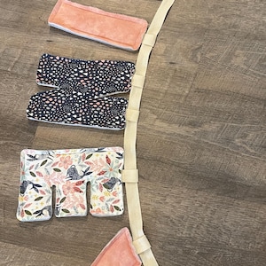 以下が含まれることがあります： さまざまな模様と色でパッドが入ったセクションがいくつかあるベビーキャリア。セクションには、フローラル、白とピンクのアクセントが付いたネイビー、ピーチ色のぬいぐるみなどがあります。クリーム色のストラップがセクションを接続しています。