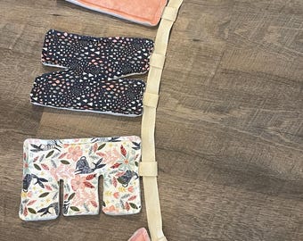 Op de afbeelding: Een draagzak met verschillende gewatteerde secties in verschillende patronen en kleuren. De secties omvatten bloemen, marineblauw met witte en roze accenten en perzikpluche. Een crèmekleurige band verbindt de secties.