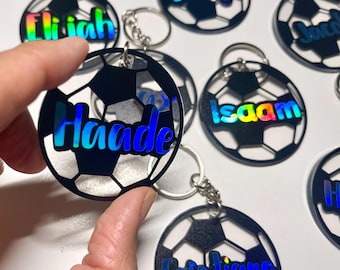 Può includere: Un set di portachiavi a forma di pallone da calcio bianco e nero con nomi iridescenti su ciascuno. I nomi sono: Elijah, George, Jack, Haade, Isaam, Cristiano.