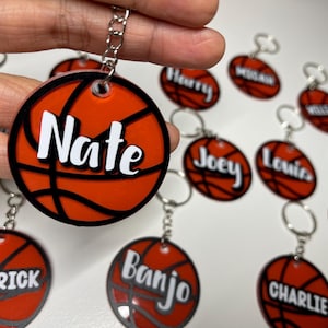 May include: A set of ten orange and black basketball-shaped keyrings with white lettering. Each keyring has a different name on it, including Nate, Kendrick, Banjo, Charlie, Joey, Harry, Louis, Ryan, and two others.