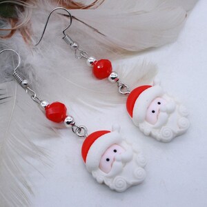May include: A pair of silver dangle earrings with red beads and white Santa Claus charms. The earrings are hanging from white feathers.