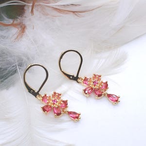 May include: A pair of gold-tone leverback earrings featuring a flower-shaped design. Each earring is adorned with multiple pink, teardrop-shaped stones arranged as petals, with a small cluster of tiny yellow stones at the center of the flower.