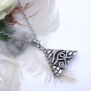 May include: A silver-toned pendant necklace with a triangular pendant featuring an ornate design. The necklace is displayed on a white surface with white feathers and a white rose in the background.