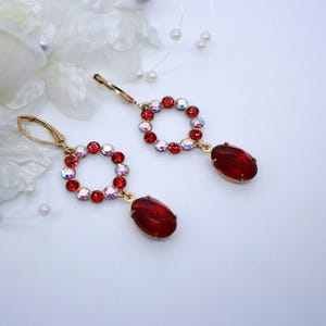 May include: A pair of gold-toned drop earrings. Each earring features a circular design with red and iridescent crystals, and a large, oval, red gemstone pendant. The earrings are displayed on a white surface with white floral accents.