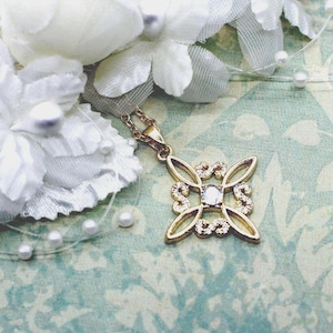 May include: A gold-toned pendant necklace featuring a symmetrical, open-work geometric design with four curved arms. The center of the pendant holds a single clear stone, while the four arms are accented with small, sparkling white crystals.