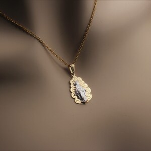 K&ouml;nnte beinhalten: Eine goldene Kettenkette mit einem Anh&auml;nger, der ein wei&szlig;es Bild der Jungfrau Maria zeigt, umgeben von einem goldenen Filigranmuster.