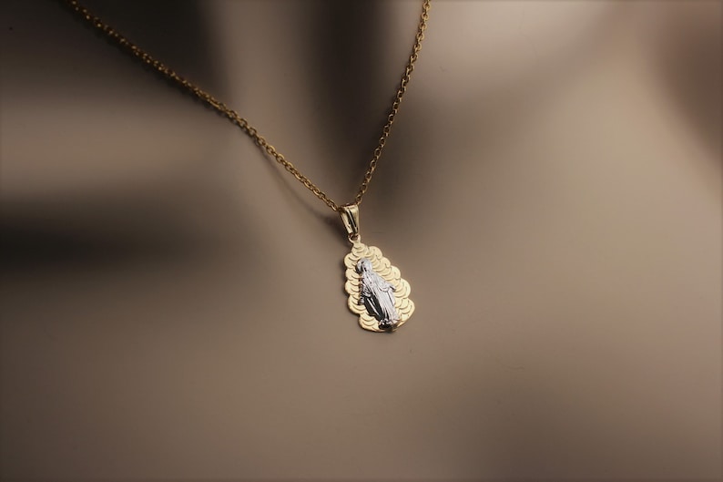 K&ouml;nnte beinhalten: Eine goldene Kettenkette mit einem Anh&auml;nger, der ein wei&szlig;es Bild der Jungfrau Maria zeigt, umgeben von einem goldenen Filigranmuster.