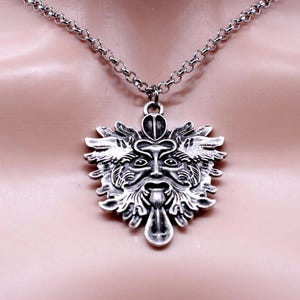 May include: A silver-colored pendant necklace featuring a detailed Green Man face design. The pendant is suspended from a silver chain. The Green Man has a beard, mustache, and leafy details around the face.