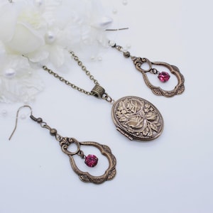 Puede incluir: Un conjunto de joyas de bronce antiguo con un collar con medallón floral y pendientes a juego. El medallón ovalado y los colgantes de los pendientes tienen un diseño de filigrana decorativo y una pequeña gema rosa redonda. Las joyas están sobre un fondo blanco.