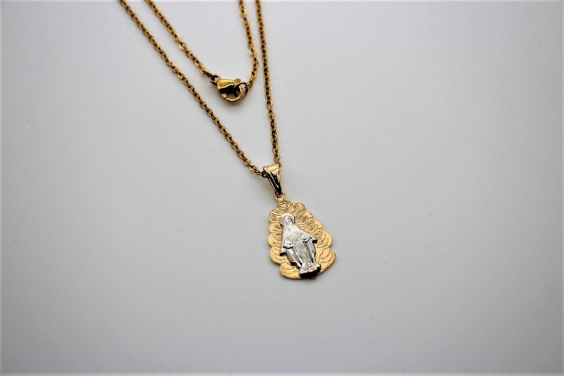 K&ouml;nnte beinhalten: Eine goldene Kettenkette mit einem Anh&auml;nger, der ein silbernes Bild der Jungfrau Maria zeigt. Der Anh&auml;nger ist von einem goldenen Design umgeben.