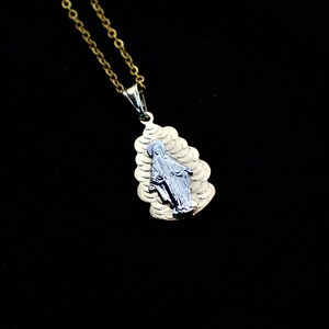 K&ouml;nnte beinhalten: Eine goldene Kettenkette mit einem Anh&auml;nger, der ein silbernes Bild der Jungfrau Maria zeigt. Der Anh&auml;nger ist tropfenf&ouml;rmig und hat ein strukturiertes Design.