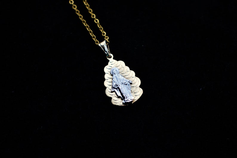 K&ouml;nnte beinhalten: Eine goldene Kettenkette mit einem Anh&auml;nger, der ein silbernes Bild der Jungfrau Maria zeigt. Der Anh&auml;nger ist tropfenf&ouml;rmig und hat ein strukturiertes Design.