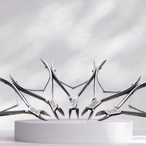 May include: A collection of five silver-toned metal cuticle nippers arranged in a fan shape. Each tool features sharp, pointed blades, a spring-loaded hinge mechanism, and textured handles for grip, with small engraved text visible on the joints.