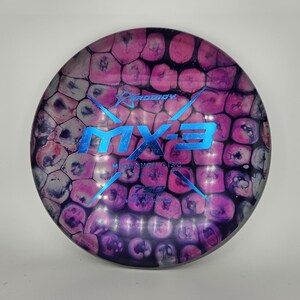 May include: A pink and purple disc golf disc with a blue and silver "MX-3" logo. The disc has a textured, honeycomb-like pattern. The text "MIDRANGE DISC" is printed on the disc.