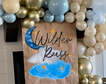 May include: A brown paper sign with a blue cloud and a crescent moon painted on it. The sign says "Wilder Russ" in black cursive writing. The sign is decorated with a garland of blue, white, and gold balloons.