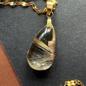 May include: A gold-colored necklace with a teardrop-shaped pendant. The pendant is clear with golden, hair-like inclusions and a gold-colored bail. The chain is also gold-colored and has a delicate, linked design. The necklace is displayed on a dark surface.