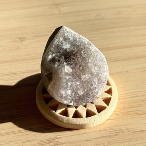 Peut inclure: Une géode de cristal en forme de goutte, de couleur gris clair, repose sur un petit support rond en bois avec un motif géométrique en forme d'étoile. La géode a une surface rugueuse et texturée.