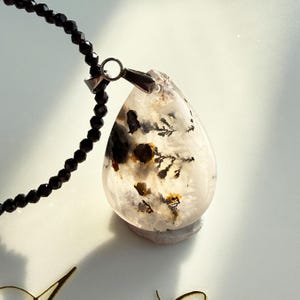 Peut inclure: Un pendentif clair en forme de goutte d'eau avec des inclusions noires et jaunes, suspendu à un collier de perles noires. Le pendentif a une bélière de couleur argentée. Le collier et le pendentif sont sur une surface blanche.
