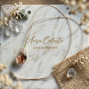 Könnte beinhalten: Aura Celeste Halskette mit einem transparenten, tropfenförmigen Anhänger mit goldenen Einschlüssen, an einer dünnen, hellbraunen Kordel. Eine kleine grüne Perle akzentuiert den Anhänger. Die Halskette liegt auf einer Marmoroberfläche mit dem Text "Aura Celeste Love & Moment".