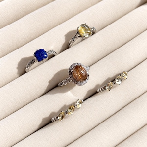 Puede incluir: Cinco anillos de tono plateado con varias piedras preciosas. Un anillo tiene una piedra cuadrada azul, uno una piedra rutilada clara, uno una gran piedra rutilada ovalada con un halo de piedras claras, y dos anillos tienen tres pequeñas piedras amarillas.