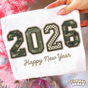 May include: White fabric with the numbers "2026" in a collegiate style, featuring camouflage and checkered patterns. The words "Happy New Year" are scripted below the numbers. The fabric is held by a hand.