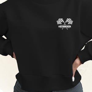 May include: Black sweatshirt featuring a white graphic of two crossed checkered racing flags. The text "LASTNAMEHERE" is printed below the flags. The sweatshirt is a crew neck style.