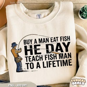 May include: A cream-coloured sweatshirt with a fishing-themed graphic. The design features a fisherman, fishing rod, and the text "Buy a man eat fish, he day, teach fish man to a lifetime." The sweatshirt has a crew neck.