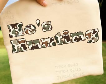 May include: A beige cushion featuring the text "He's Hunting" in a camouflage design. The cushion is held by a hand, with blurred green grass in the background. The words "Typical Deals" are printed on the cushion.