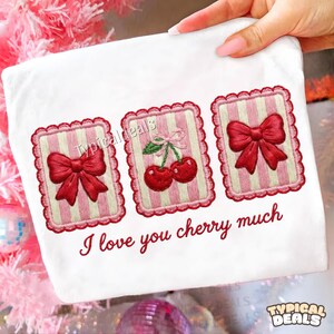 Puede incluir: Tela blanca con diseños bordados de cerezas y lazos. El diseño incluye tres marcos rectangulares con fondos a rayas rosas y blancas. Los marcos tienen lazos rojos y cerezas. El texto "I love you cherry much" está debajo.