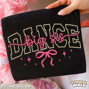 May include: A black t-shirt featuring the word DANCE in large beige block letters. Overlaid is the text big sis in pink cursive script with small hearts, a pink bow graphic, and pink star accents printed on the front of the garment.
