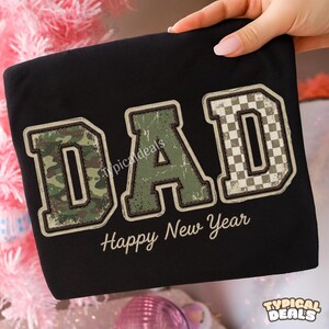 Könnte beinhalten: Schwarzes Sweatshirt mit dem Wort "DAD" in einer verblassten Schriftart im College-Stil. Der Buchstabe "D" hat ein Camouflage-Muster, das "A" ist grün und das letzte "D" hat ein Schachbrettmuster. Der Satz "Happy New Year" steht darunter.