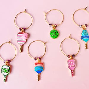 May include: A collection of gold-toned wine charms with colorful pickleball paddle designs. Each charm features a different paddle design in shades of pink, green, blue, and red, with some including the text "LET'S PLAY" or "PICKLEBALL".