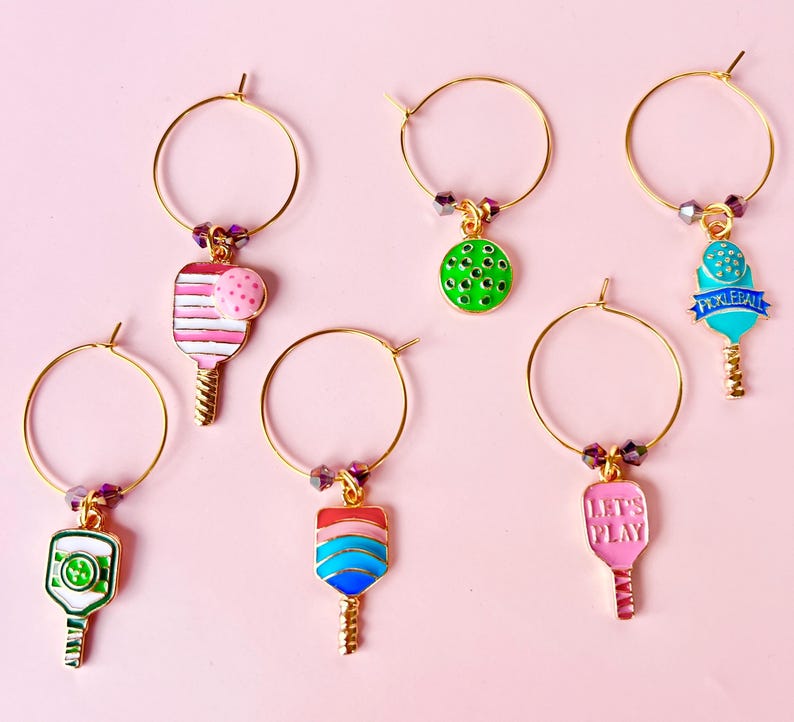 May include: A collection of gold-toned wine charms with colorful pickleball paddle designs. Each charm features a different paddle design in shades of pink, green, blue, and red, with some including the text "LET'S PLAY" or "PICKLEBALL".