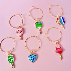 May include: Gold-tone hoop earrings with colorful pickleball paddle charms. The charms feature various designs, including a heart with blue dots, a paddle with the text "Pickeball", and paddles with green, red, and blue accents. The earrings are set against a pink background.