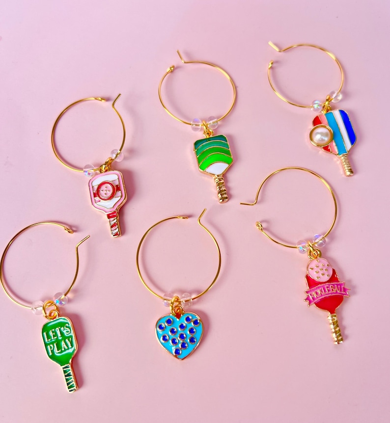 May include: Gold-tone hoop earrings with colorful pickleball paddle charms. The charms feature various designs, including a heart with blue dots, a paddle with the text "Pickeball", and paddles with green, red, and blue accents. The earrings are set against a pink background.