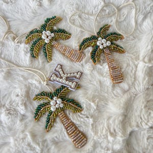 Op de afbeelding: Vier kralen palmboomornamenten met wit touw om op te hangen. De palmbladeren zijn groen en goud, met witte parelaccenten. De stammen zijn bruin en goud. E&eacute;n ornament heeft een witte letter "K" met parelaccenten.