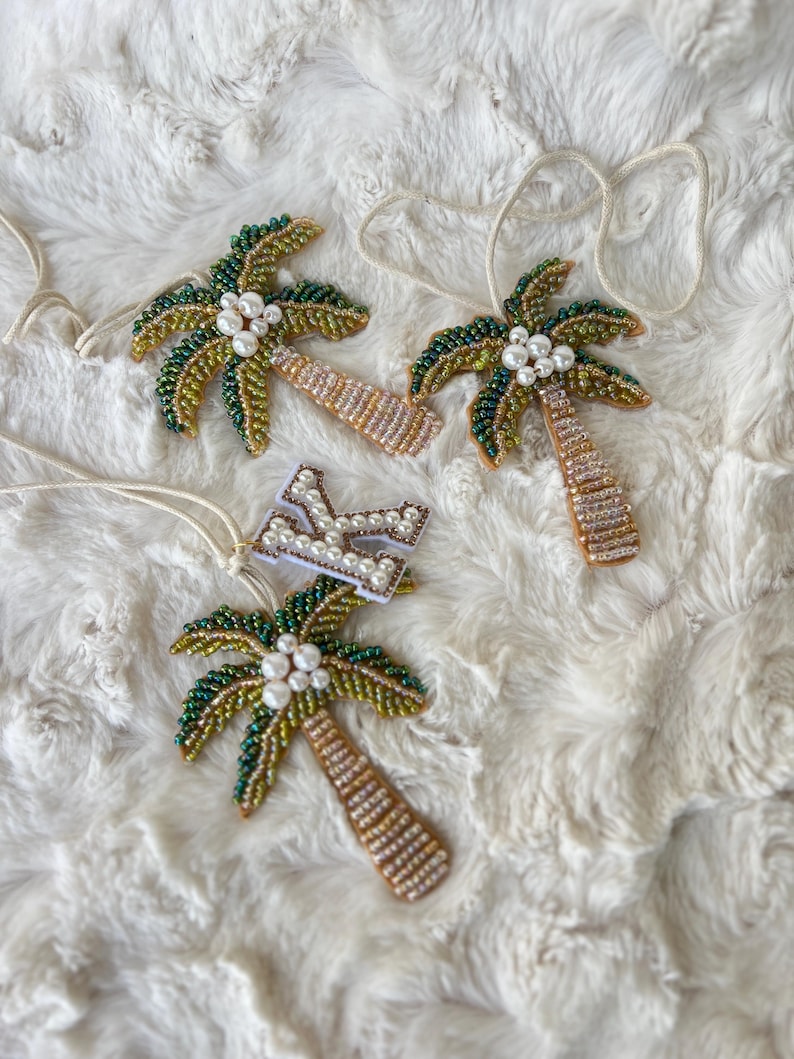 Op de afbeelding: Vier kralen palmboomornamenten met wit touw om op te hangen. De palmbladeren zijn groen en goud, met witte parelaccenten. De stammen zijn bruin en goud. E&eacute;n ornament heeft een witte letter "K" met parelaccenten.
